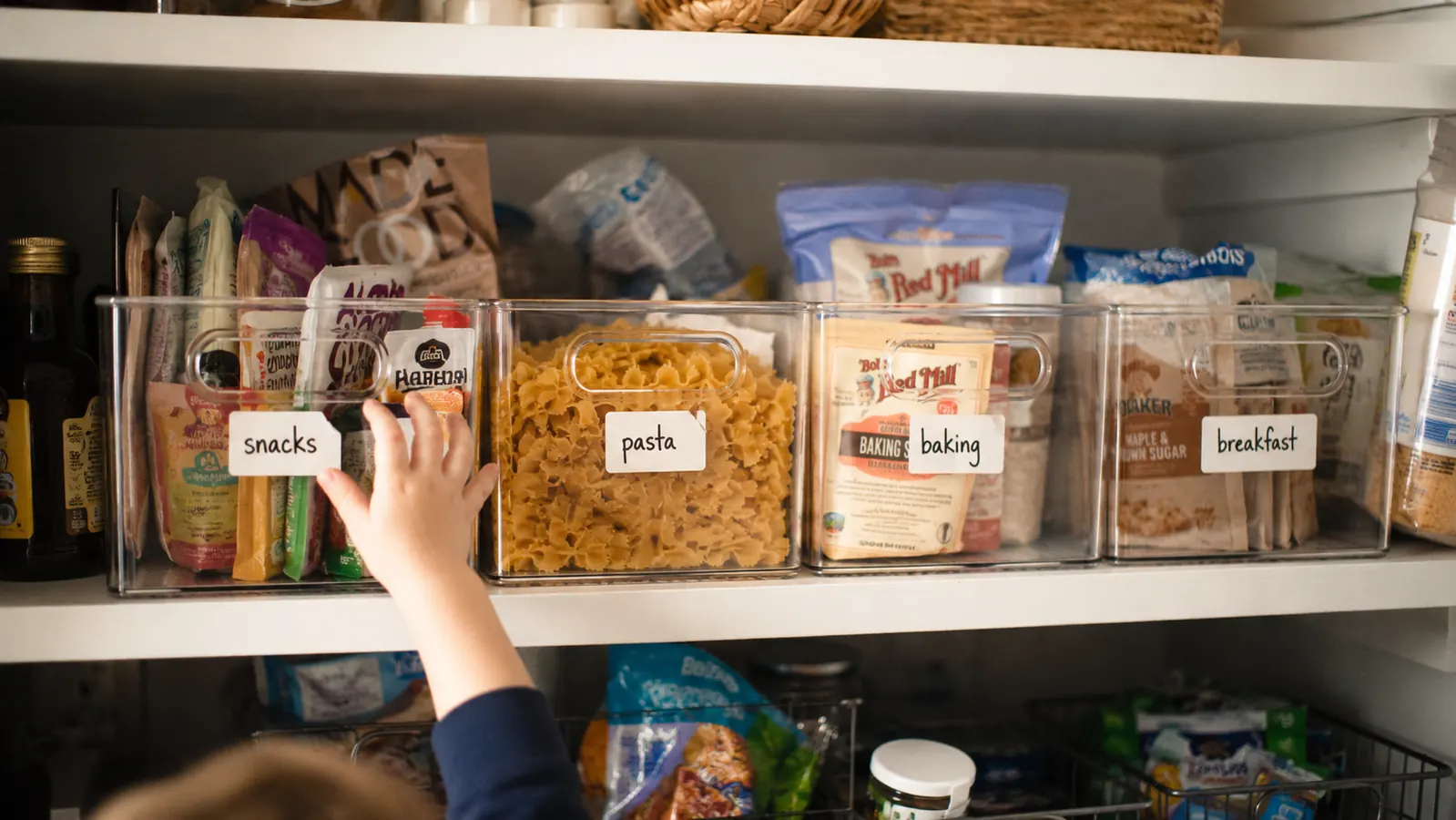 Pantry shelf with clear bins organized by food category, labels visible on a few, a child's hand reaching into the snacks bin